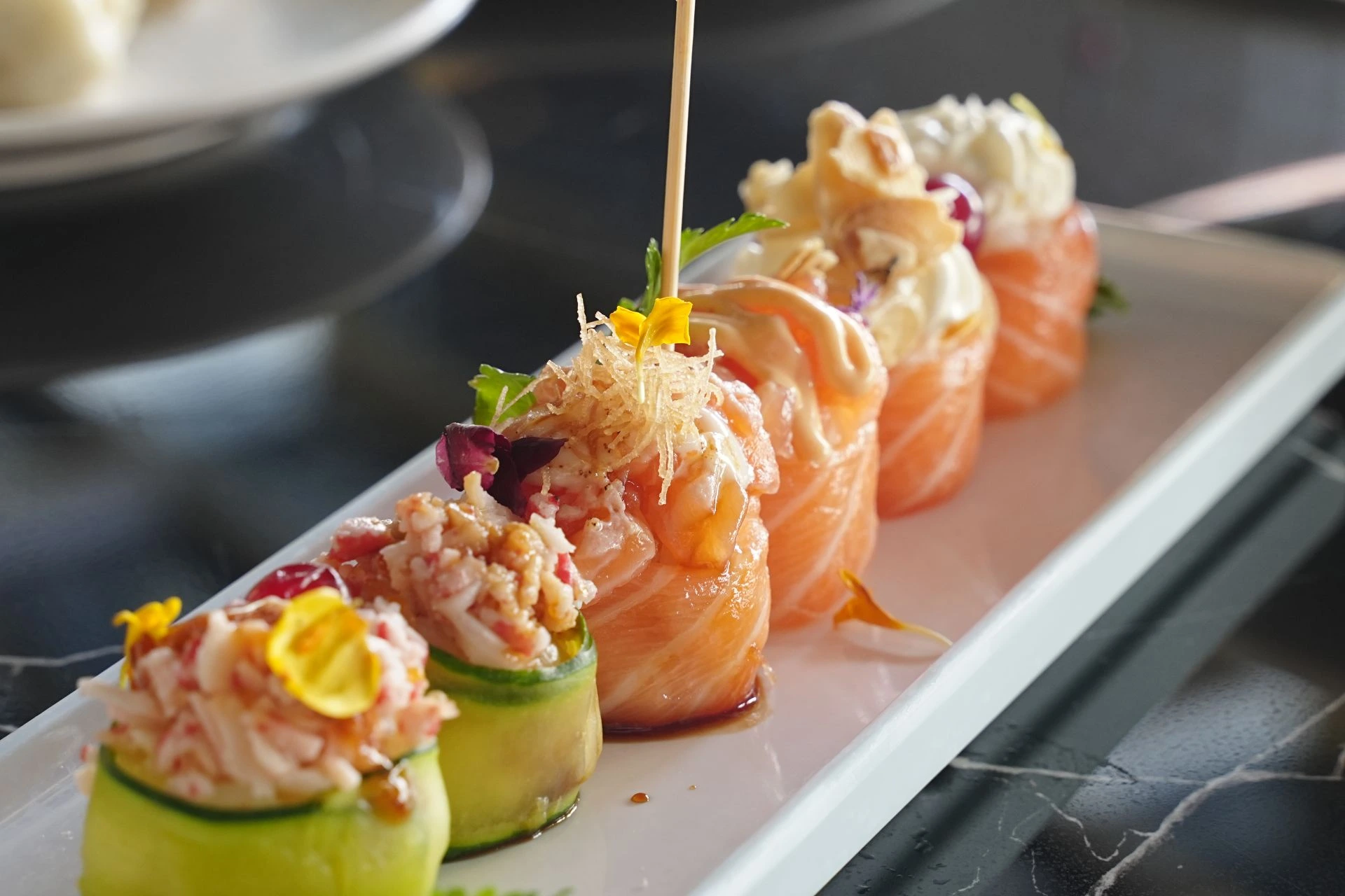 Su un vassoio lungo bianco sono disposti ordinatamente piatti giapponesi raffinati, tra cui rotoli di cetriolo (con ripieno di carne di granchio tritata) e rotoli di salmone, decorati con petali gialli, frammenti croccanti fatti in padella e salsa, con spiedini di legno inserti, il tutto presentato in modo elegante.