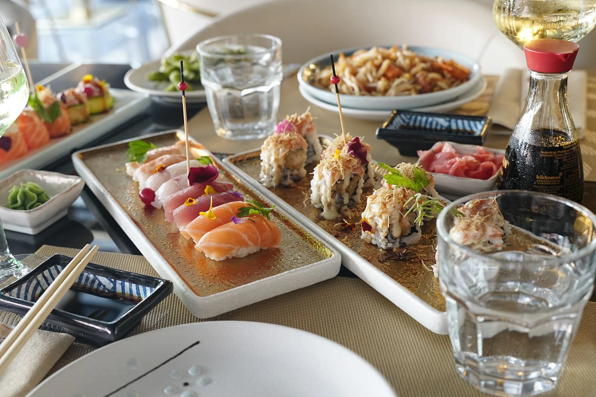 Un tavolo ricoperto di cucina giapponese, con sushi di salmone, tonno e altri pesci crudi su vassoi dorati, nonché rotoli di sushi fritti, abbinati a wasabi, salsa di soia (marca KIKKOMAN), calici di vino bianco, bicchieri d'acqua e bacchette. Sullo sfondo sono visibili altri piatti, presentando una scena ricca e raffinata di cibi giapponesi.